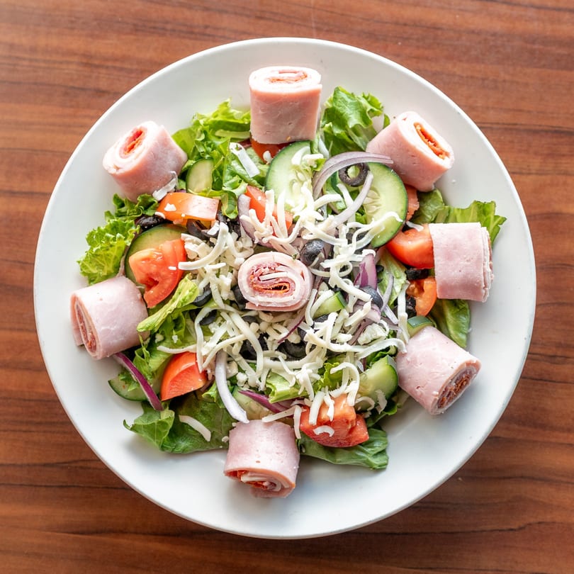 A fresh salad with lettuce, tomatoes, cucumbers, olives, onions, cheese, and ham rolls.